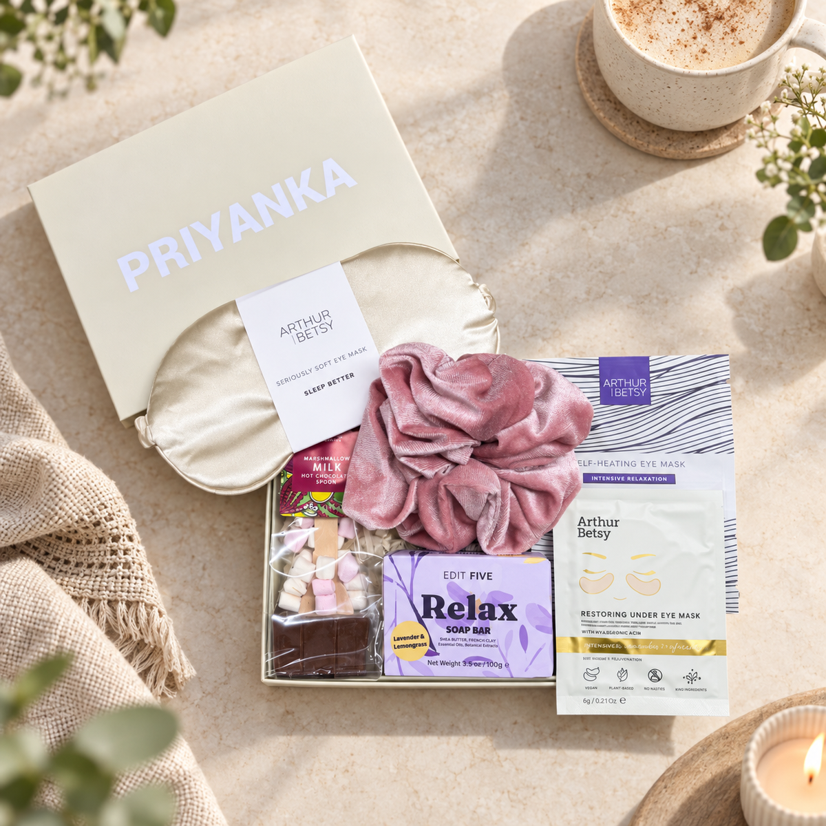 A curated gift set from Arthur Betsey and Priyanka, beautifully arranged in a flat lay. The set includes a cream satin sleep mask, a dusty pink silk scrunchie, a purple 'Relax' hair mask, an eye mask packet, and a chocolate bar, all presented in a cream-colored gift box with the brand names clearly visible. Hallby letterbox gift for her with Relax soap, warming eye mask, satin eye mask and hot chocolate