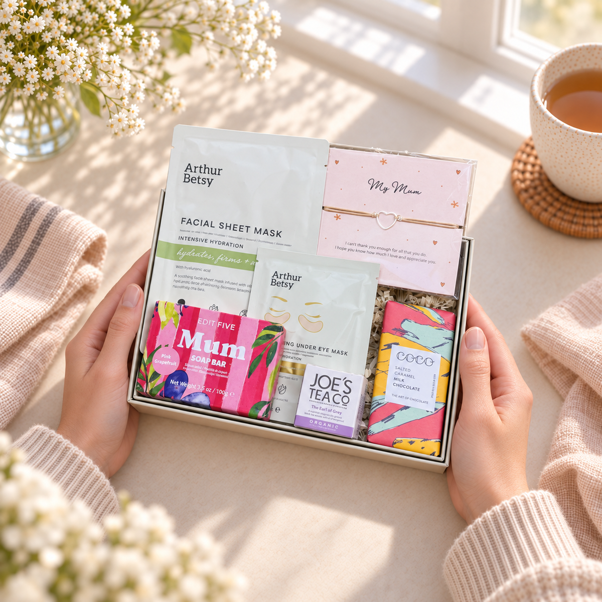 A gift box of skincare and tea products held by two hands on a cozy windowsill. The box contains Arthur Betsy facial sheet masks, a pink 'My Mum' gift card, a bag of Joe's Tea Teacake, and a colorful box of Oreo chocolate. Mother’s Day letterbox gift with self-care treats, Joe’s Tea, chocolate and a meaningful string bracelet