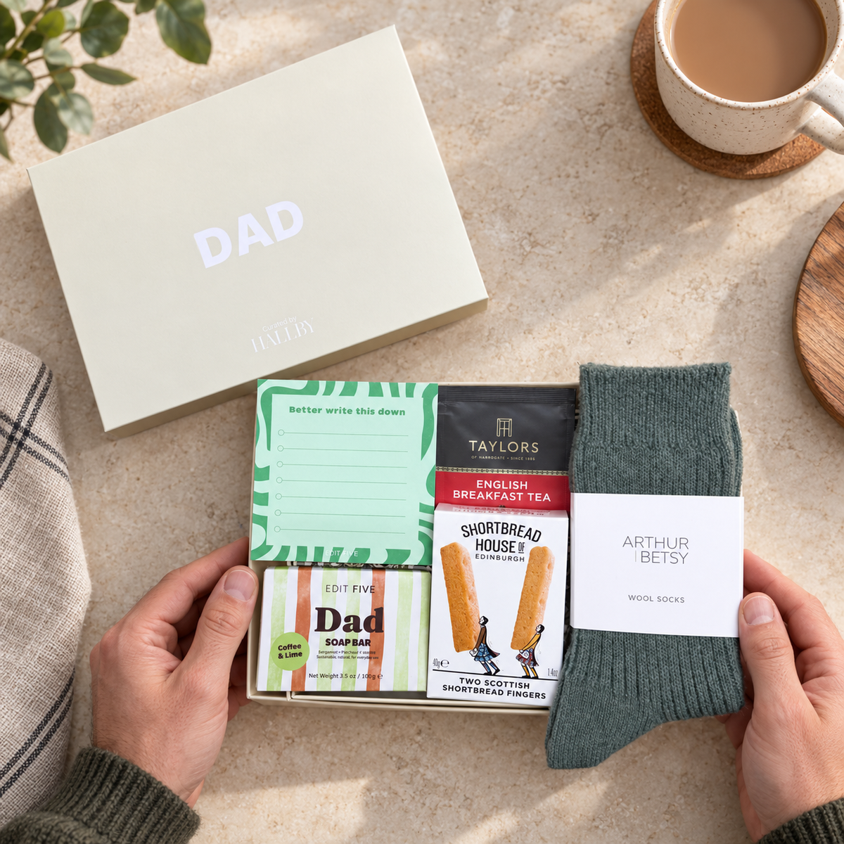 A curated gift set for Dad laid out on a light stone surface, held by two hands. The set includes a cream gift box labeled DAD, a green patterned note card, a black and red Taylors English Breakfast Tea packet, a Shortbread House of Edinburgh biscuit packet, a packet of dad-themed shortbread papers, and a pair of dark green Arthur Betsy wool socks with a white label. Hallby letterbox gift for Dad with Twilight wool socks, Dad soap, shortbread, English Breakfast tea and a Better Write This Down memo pad
