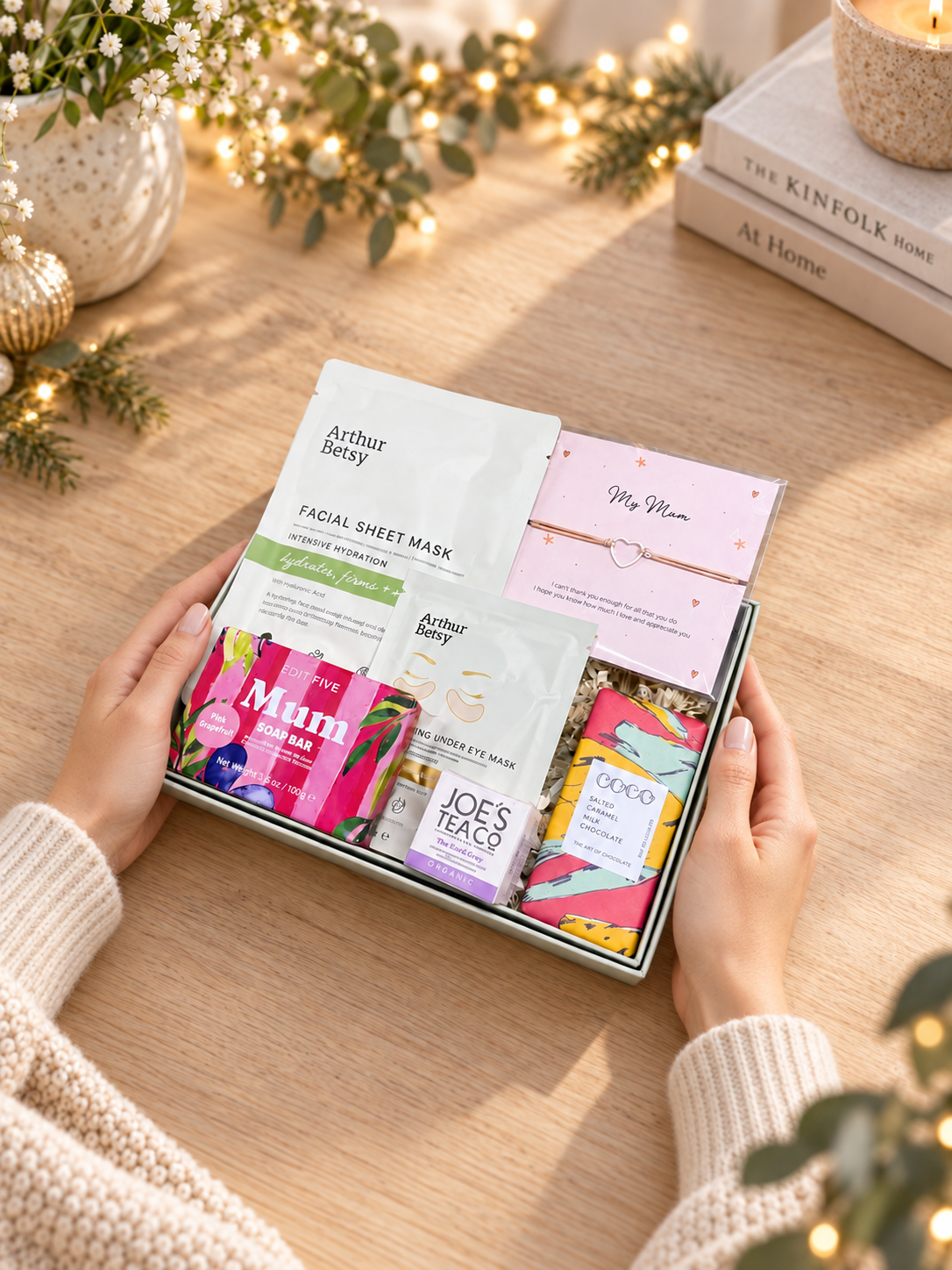 A gift box of facial sheet masks held by hands over a wooden table decorated with fairy lights and books. Christmas gift for Mum with skincare, tea, salted caramel chocolate, artisan soap and My Mum bracelet
