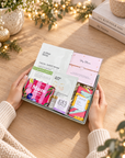 A gift box of facial sheet masks held by hands over a wooden table decorated with fairy lights and books. Christmas gift for Mum with skincare, tea, salted caramel chocolate, artisan soap and My Mum bracelet
