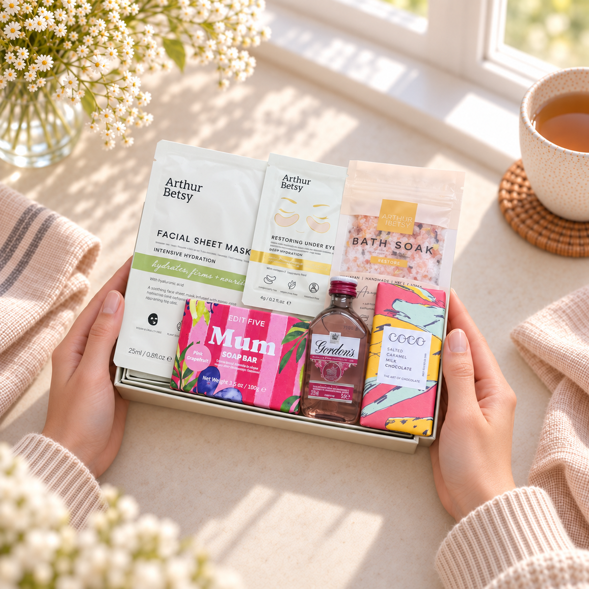 A gift box held by two hands on a windowsill, filled with skincare and beauty products including Arthur Betsy facial sheet mask, redosing eye gel, bath soap, and a small bottle of Tenders tea, surrounded by cozy props like a cup of tea and flowers. Relaxing gifts for Mum featuring Arthur Betsy Restore Bath Soak, gold under-eye patches and Gordon’s Pink Gin