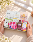 A gift box held by two hands on a windowsill, filled with skincare and beauty products including Arthur Betsy facial sheet mask, redosing eye gel, bath soap, and a small bottle of Tenders tea, surrounded by cozy props like a cup of tea and flowers. Relaxing gifts for Mum featuring Arthur Betsy Restore Bath Soak, gold under-eye patches and Gordon’s Pink Gin