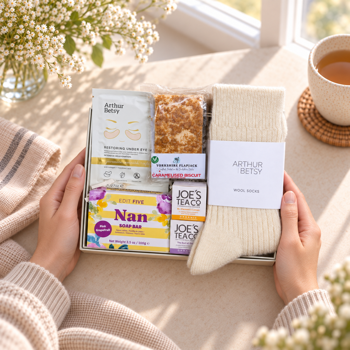 A gift box held by two hands, featuring Arthur Betsy wool socks, a chocolate biscuit, a sachet of soothing eye cream, and Joe's Tea Co. tea bars, styled on a warm beige table with a cup of tea and flowers in the background.