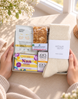 A gift box held by two hands, featuring Arthur Betsy wool socks, a chocolate biscuit, a sachet of soothing eye cream, and Joe's Tea Co. tea bars, styled on a warm beige table with a cup of tea and flowers in the background.