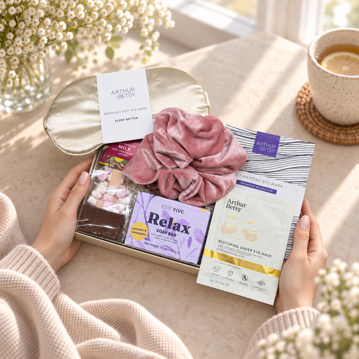 A gift box held by two hands in a cozy indoor setting, featuring Arthur Betty skincare items including a pink velvet scrunchie, a Relax cold pack, and an eye mask packet, with a cream eye mask box behind it. 
Relaxing Christmas gift for her featuring Arthur Betsy gold under-eye patches and cosy self-care treats
