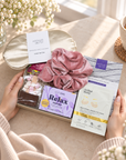 A gift box held by two hands in a cozy indoor setting, featuring Arthur Betty skincare items including a pink velvet scrunchie, a Relax cold pack, and an eye mask packet, with a cream eye mask box behind it. 
Relaxing Christmas gift for her featuring Arthur Betsy gold under-eye patches and cosy self-care treats
