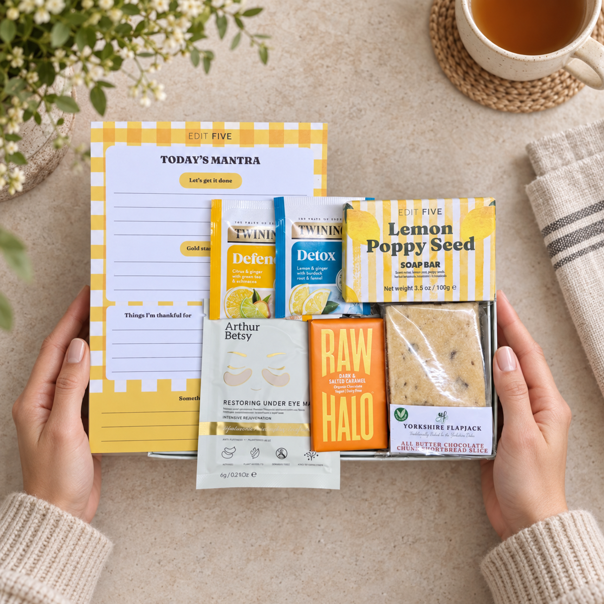 A styled flat-lay showing a curated gift set held by two hands. The set includes a yellow and white planner pad titled 'TODAY'S MANTRA', Twinin tea packets (Defence, Detox, and Lemon Poppy Seed), an Arthur Betty eye mask packet, a RAW HALO supplement box, and a Yorkshire Flapjack cookie. Hallby sunshine letterbox gift for her with positive notepad, bath salts, face mask, soap and chocolate