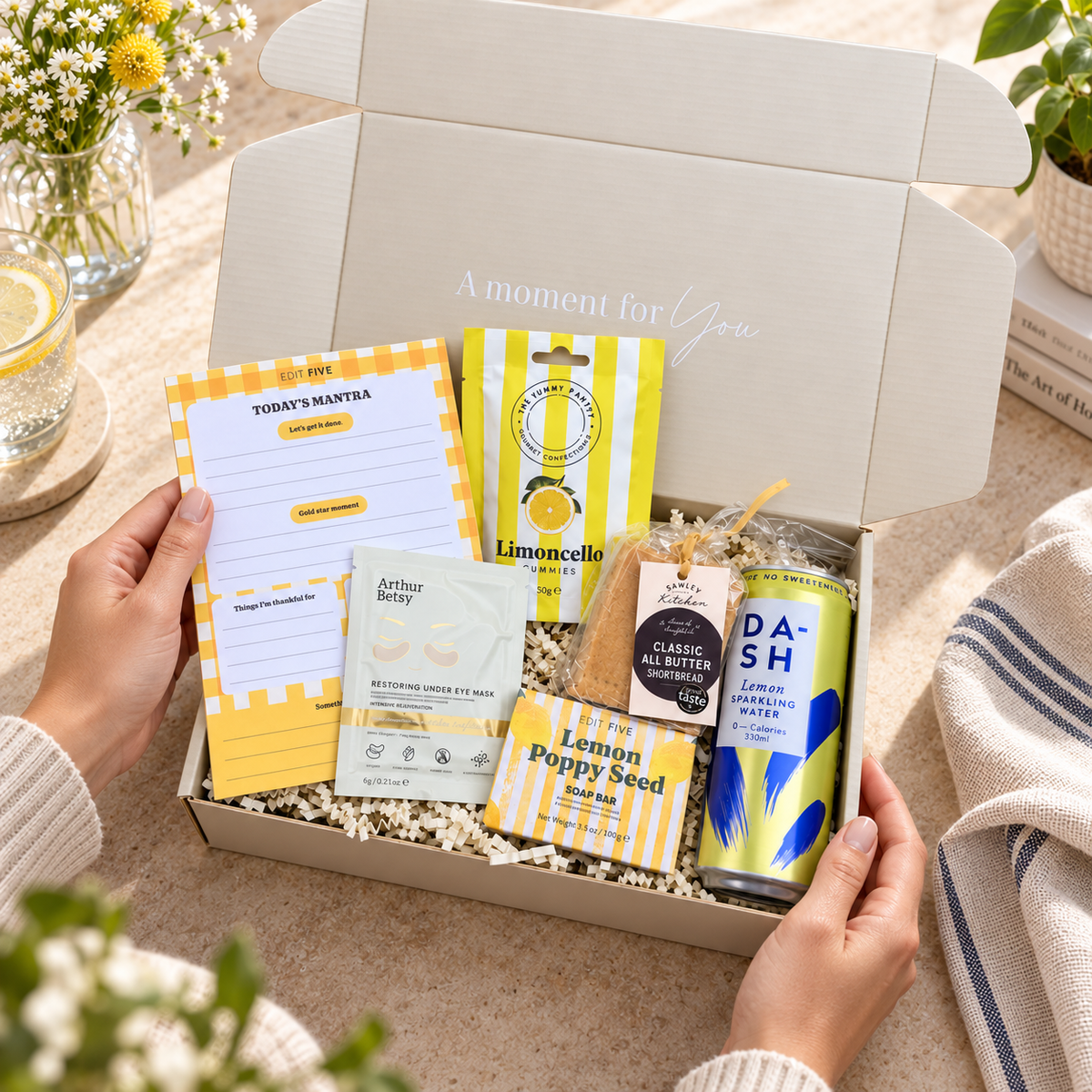 A curated gift box held by two hands, featuring various wellness and lifestyle items including a lemon candle, a notepad, a bath bomb, a seed packet, and a lemonade can, set against a warm, sunlit background with flowers and a striped towel. Hand-packed Hallby gift for her with positive stationery, refreshing lemon treats and pampering products