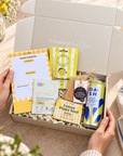 A curated gift box held by two hands, featuring various wellness and lifestyle items including a lemon candle, a notepad, a bath bomb, a seed packet, and a lemonade can, set against a warm, sunlit background with flowers and a striped towel. Hand-packed Hallby gift for her with positive stationery, refreshing lemon treats and pampering products