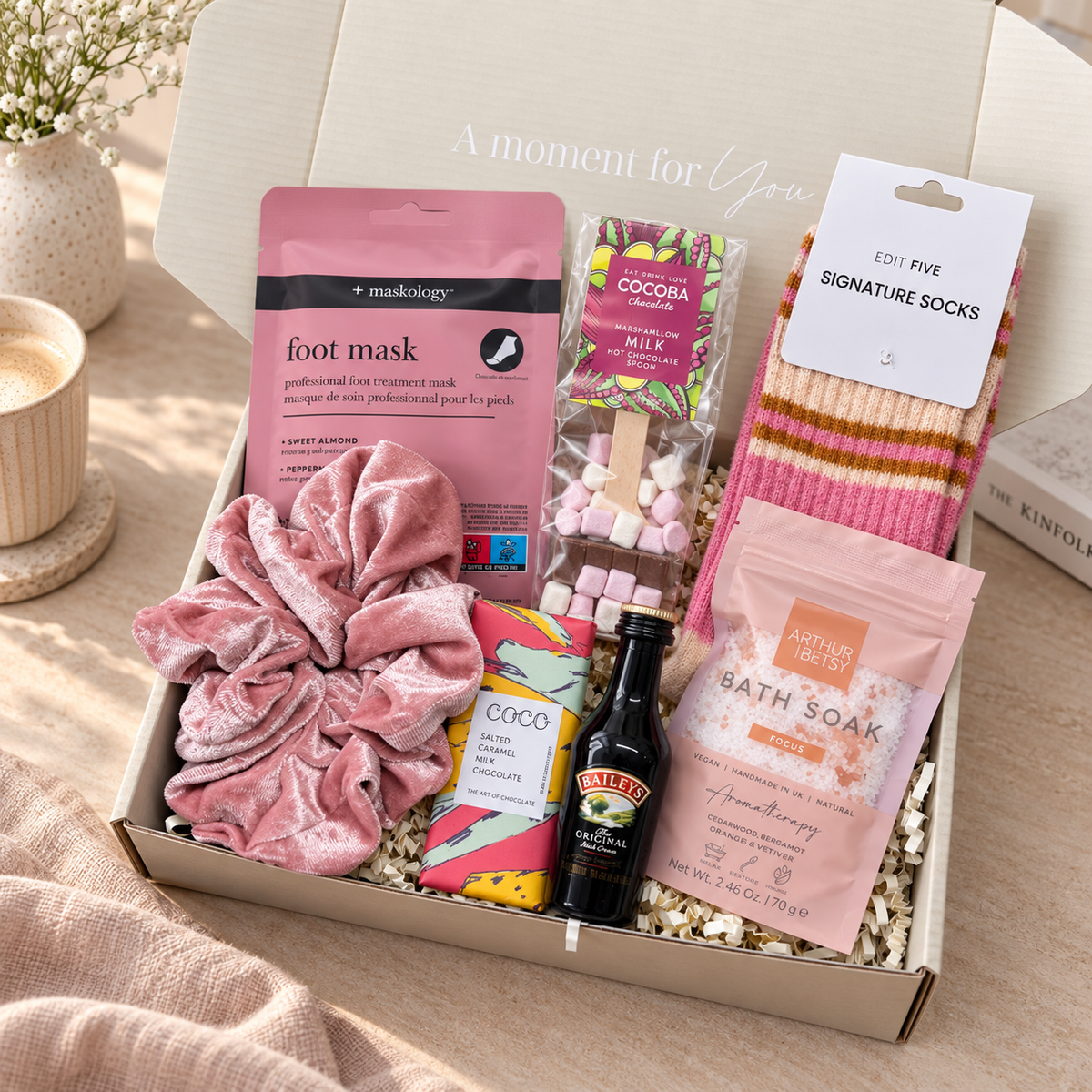 A curated gift box set in a light wood box labeled 'A moment for You', containing a pink velvet scrunchie, a pink foot mask bag by Masology, a bag of cocoa milk chocolate treats, a pack of striped socks, a bottle of Bailey's whiskey, and a pink bath soak bag, arranged on a beige fabric surface. Christmas gift box for women containing socks, Cocoba hot chocolate spoon, Maskology foot mask and Arthur Betsy bath soak