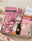 A curated gift box set in a light wood box labeled 'A moment for You', containing a pink velvet scrunchie, a pink foot mask bag by Masology, a bag of cocoa milk chocolate treats, a pack of striped socks, a bottle of Bailey's whiskey, and a pink bath soak bag, arranged on a beige fabric surface. Christmas gift box for women containing socks, Cocoba hot chocolate spoon, Maskology foot mask and Arthur Betsy bath soak