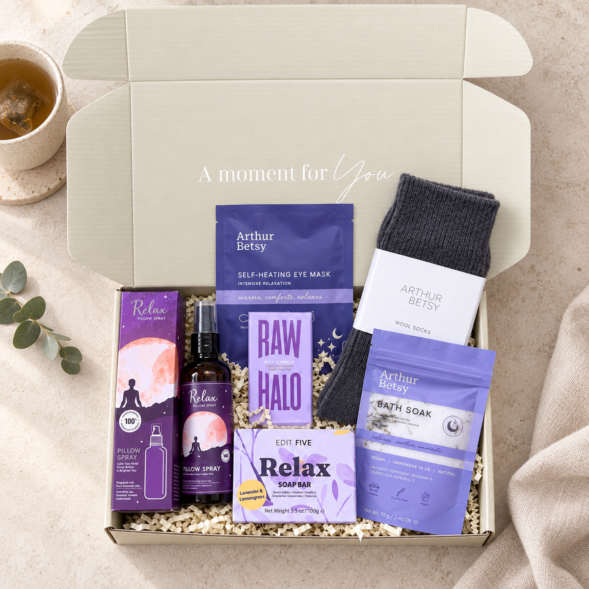 A curated self-care gift box from Arthur Betsy and Relax, containing a self-heating eye mask, eye drop bottle, soap bar, bath soap, and a dark gray sock, arranged in a kraft box with shredded paper filler on a beige fabric background. Relaxing self-care gift box with wool socks, sleep bath soak, self-heating eye mask, Relax soap, pillow spray and Raw Halo chocolate.