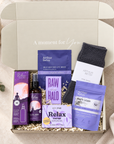A curated self-care gift box from Arthur Betsy and Relax, containing a self-heating eye mask, eye drop bottle, soap bar, bath soap, and a dark gray sock, arranged in a kraft box with shredded paper filler on a beige fabric background. Relaxing self-care gift box with wool socks, sleep bath soak, self-heating eye mask, Relax soap, pillow spray and Raw Halo chocolate.