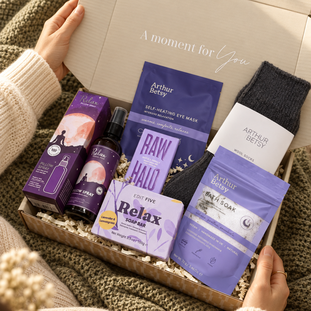 A cozy gift box set held open by hands in a knit sweater, containing Arthur Berry skincare items including a self-heating eye mask, facial serum, and mask, alongside a dark gray sock and a purple bath soak pouch. Relaxing self-care gift box with wool socks, sleep bath soak, self-heating eye mask, Relax soap, pillow spray and Raw Halo chocolate.