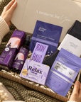 A cozy gift box set held open by hands in a knit sweater, containing Arthur Berry skincare items including a self-heating eye mask, facial serum, and mask, alongside a dark gray sock and a purple bath soak pouch. Relaxing self-care gift box with wool socks, sleep bath soak, self-heating eye mask, Relax soap, pillow spray and Raw Halo chocolate.